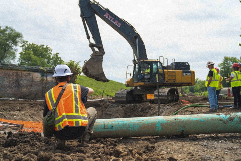 A crew member crouched beside new pipe, an Atlas excavator working the cut behind