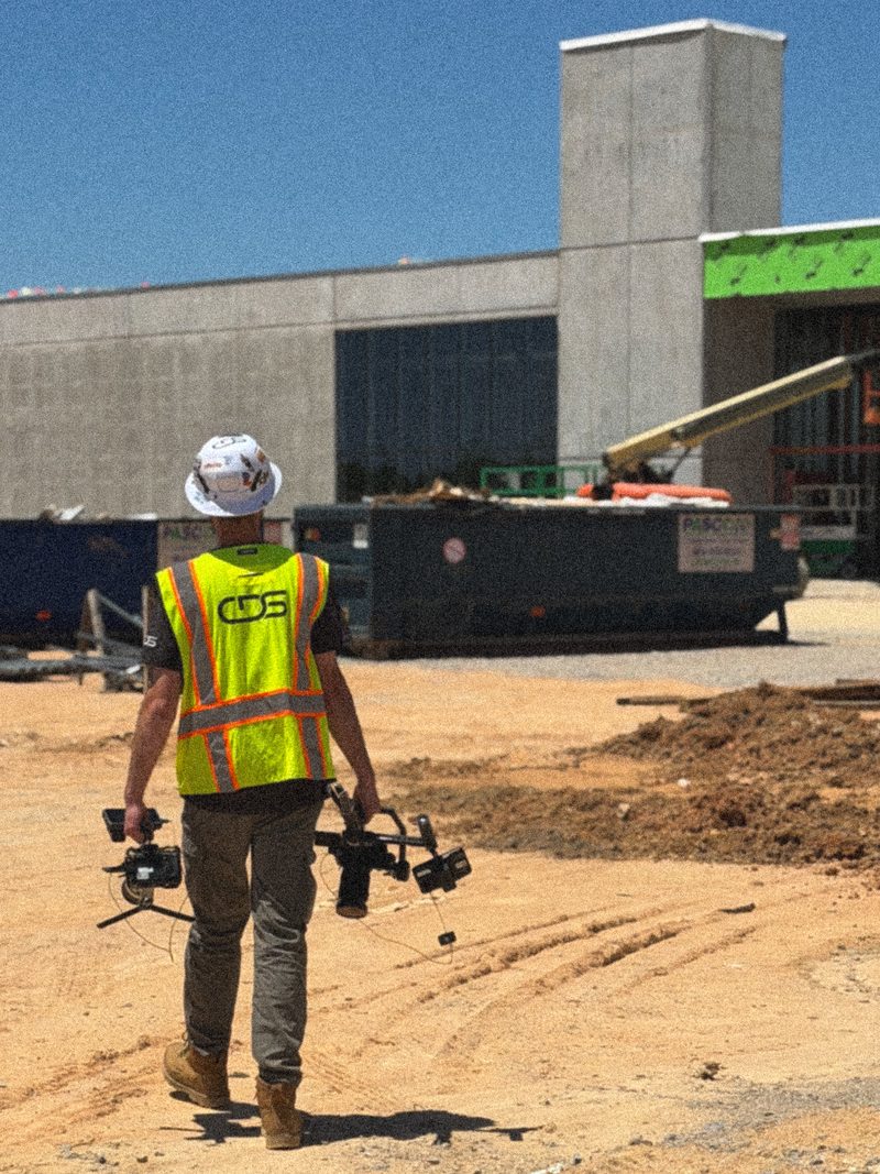 A CDS shooter in a branded vest carrying a camera rig across a graded site