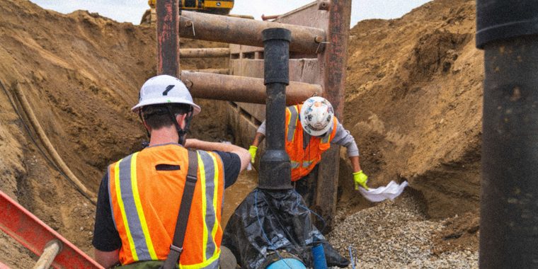 Two workers in orange high-vis setting pipe in a shored trench, an excavator on the bank above