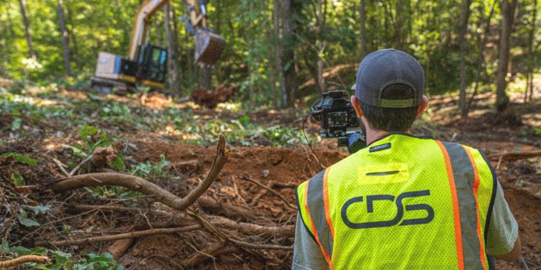 A CDS shooter in a branded high-vis vest filming an excavator clearing a wooded slope