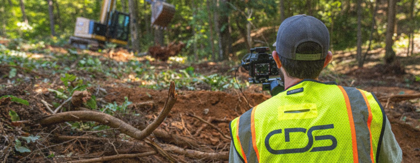 A CDS shooter in a branded high-vis vest filming an excavator clearing a wooded slope