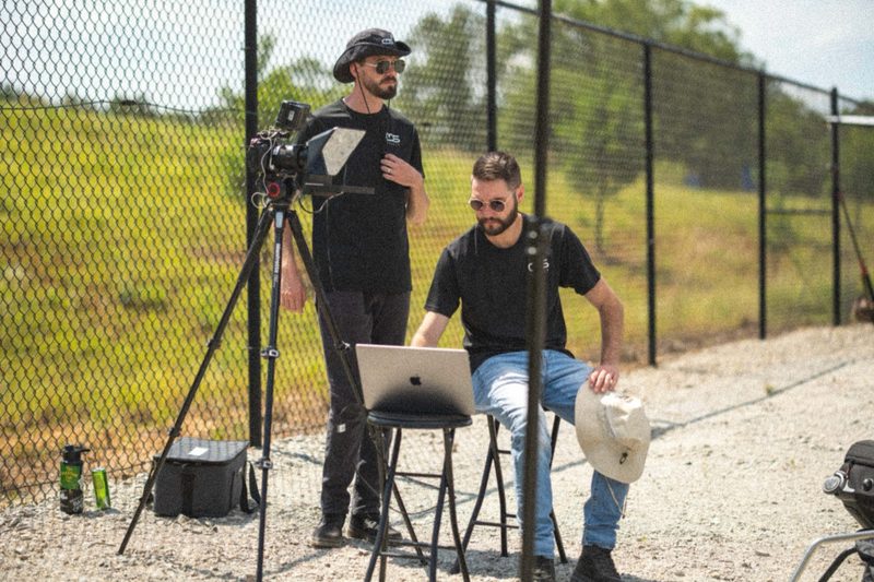 Two shooters working camera and laptop at the fence line above a green field