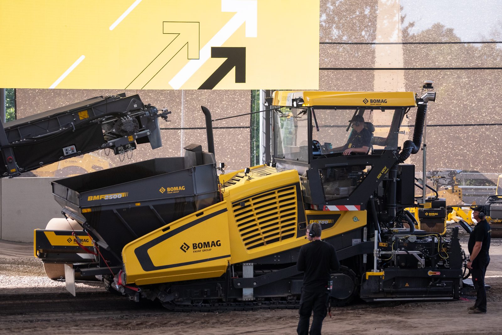 A BOMAG paver and material transfer vehicle staged side by side on a lot, an operator in the paver seat and crew standing alongside