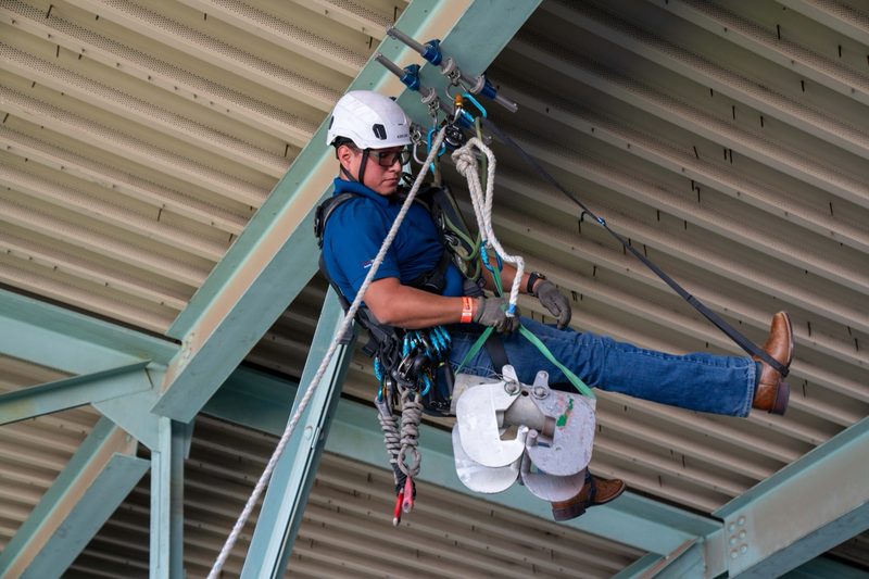 A rope-access technician suspended under the steel roof deck, working on rope