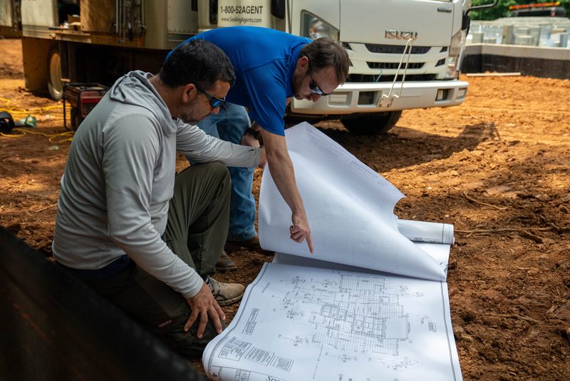 Two men crouched over the plans rolled out on the slab edge, truck behind them