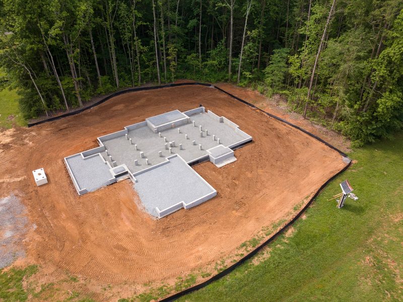 Aerial over a poured foundation on a cleared red clay lot ringed by trees