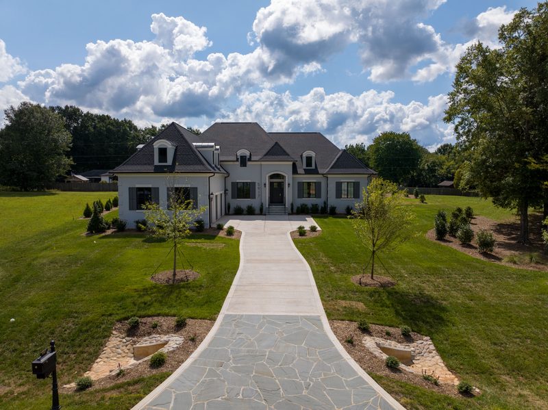 A finished custom home at the end of a stone walkway, lawn and treeline behind