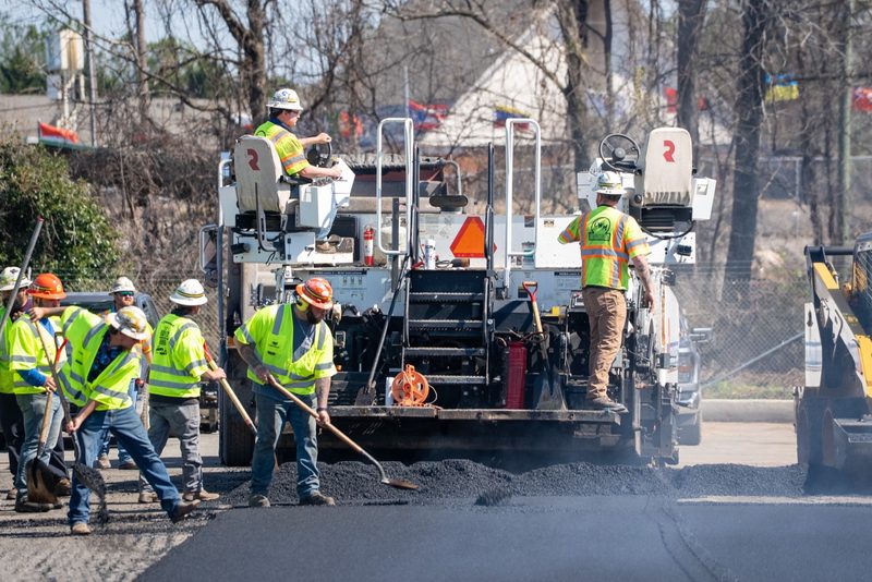 The paving crew working the mat behind the machine, rakes and shovels going
