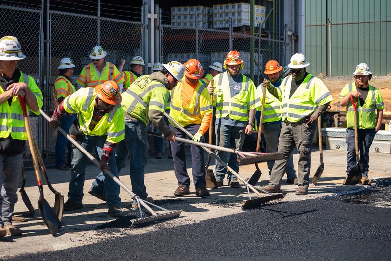 A line of crew working a fresh asphalt patch with shovels and rakes
