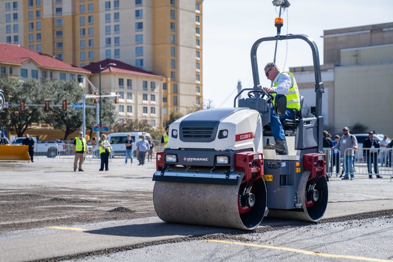 A Dynapac tandem roller finishing a lot, operator up top and a crowd along the barrier
