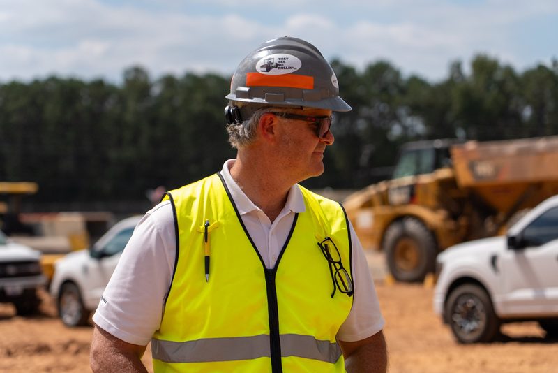 A worker in high-vis and hard hat on the jobsite, loader and pickups behind him