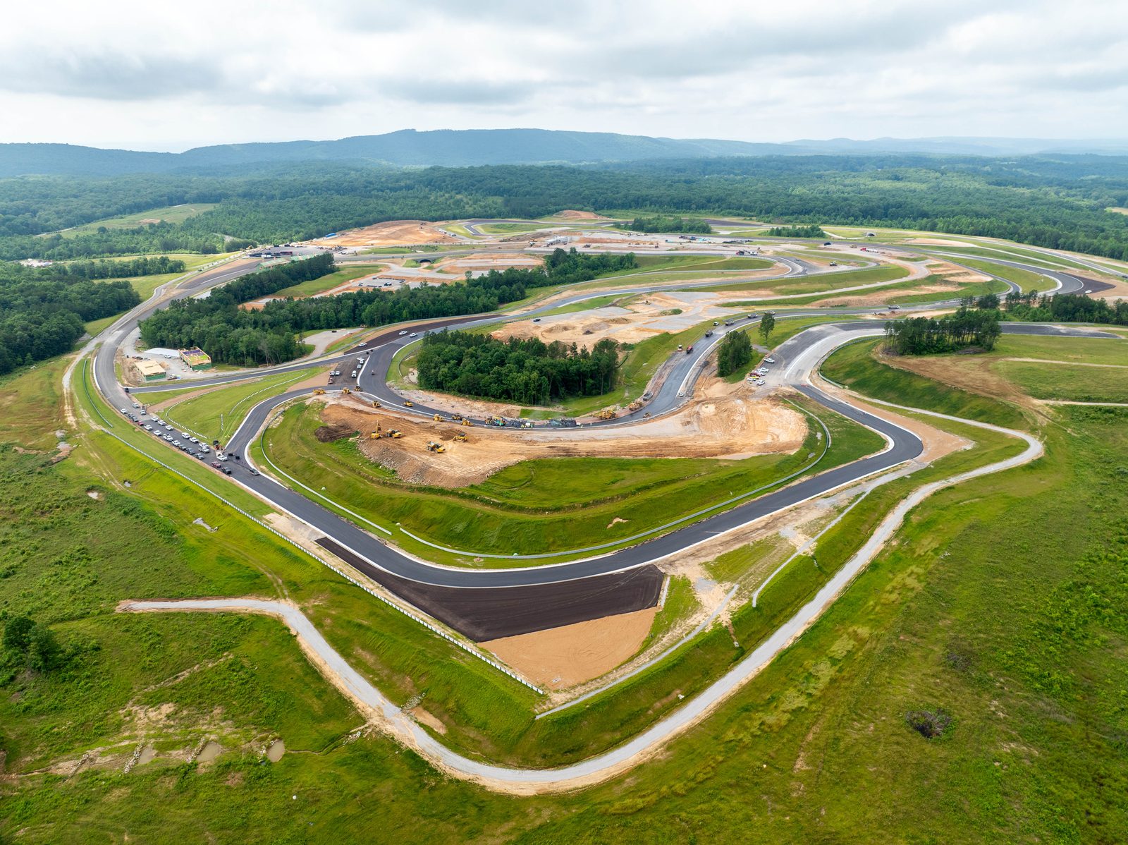 Aerial of a motorsports circuit under construction, a fresh asphalt ribbon curving through green infield and treeline
