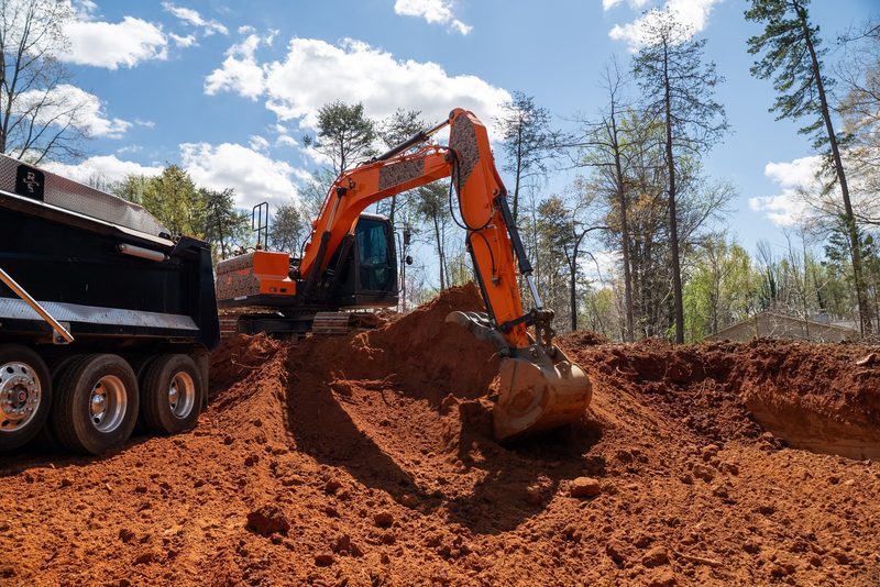 An excavator loading red dirt into the truck at the edge of the cleared timber