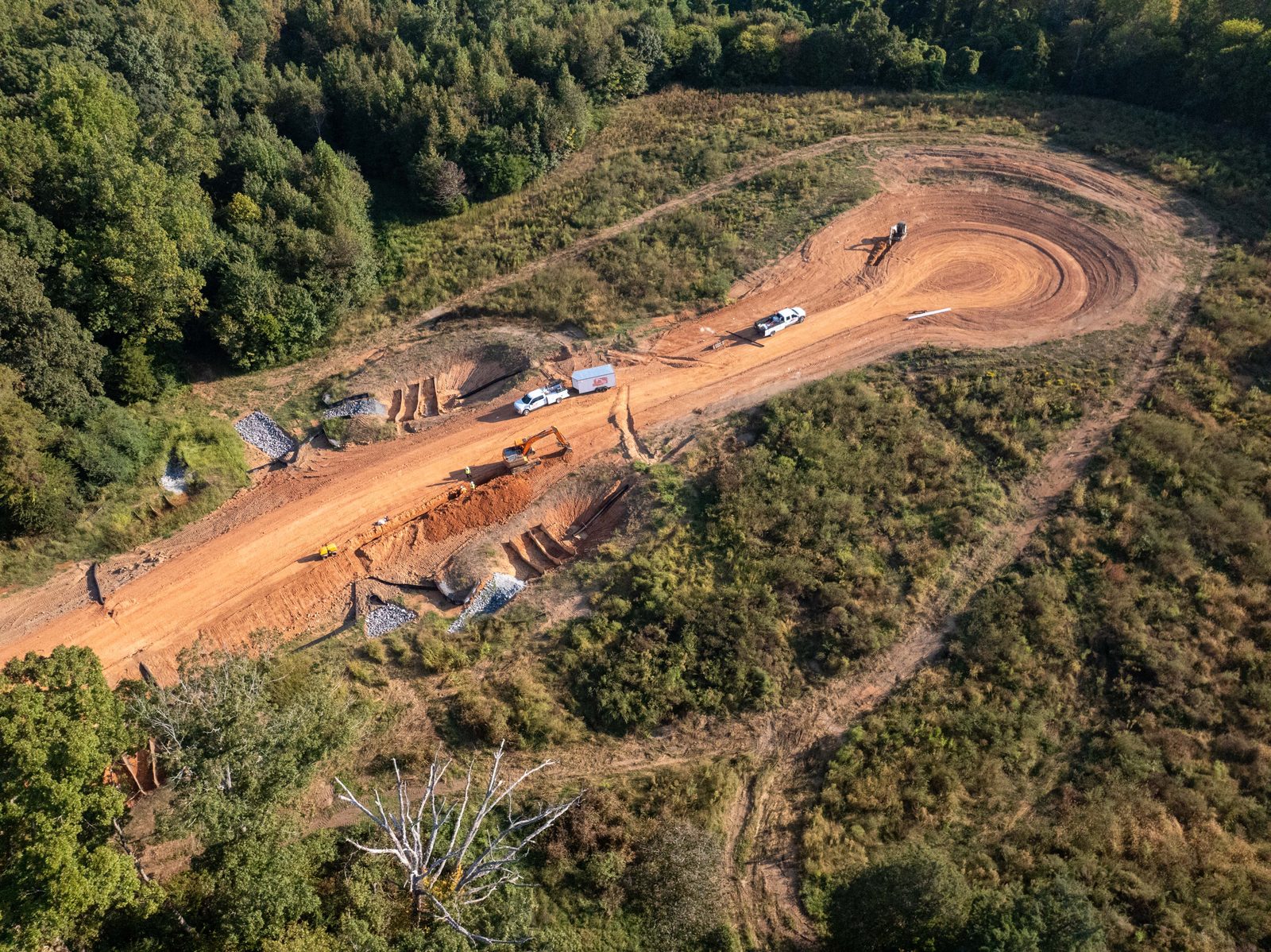 Aerial of a red-clay grading site cut into a wooded hillside, the haul road curving through it
