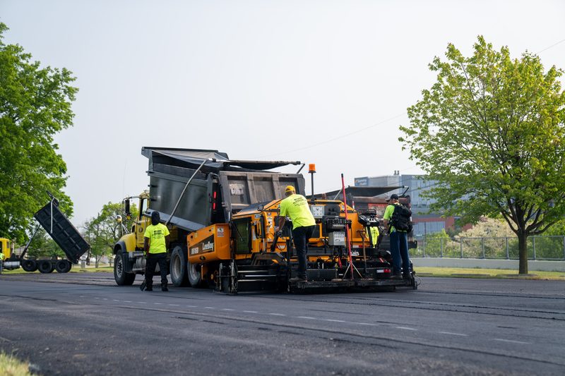 Wide on the paving train crossing the lot, truck feeding at the front