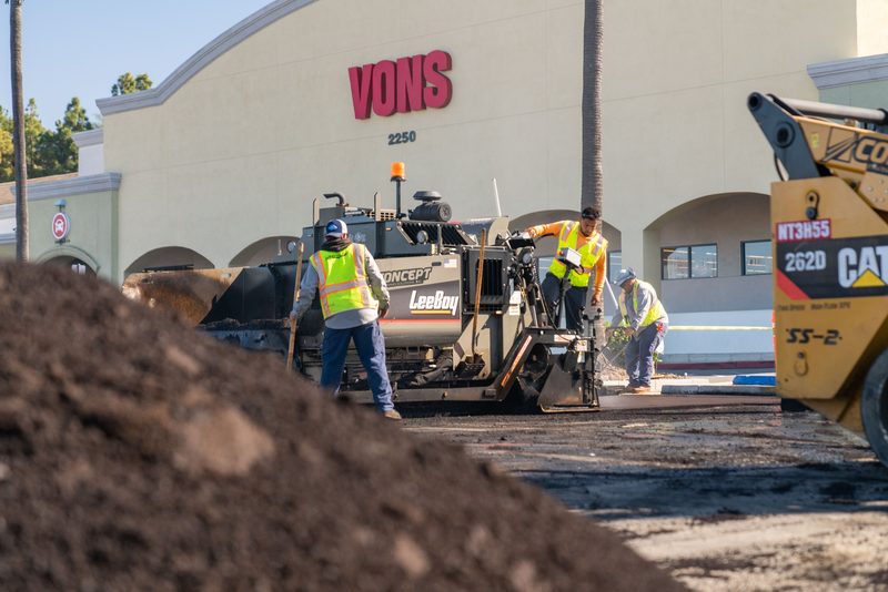 A paving crew working the storefront lot, fresh mix piled in the foreground