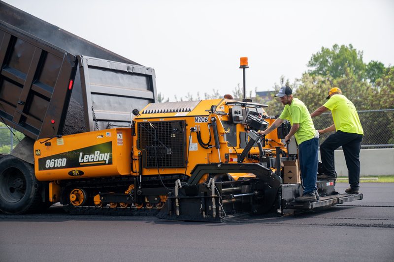 A dump truck feeding a LeeBoy 8616D paver, screed crew riding alongside