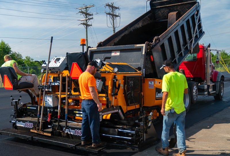 A LeeBoy paver taking a load, crew watching the hopper fill