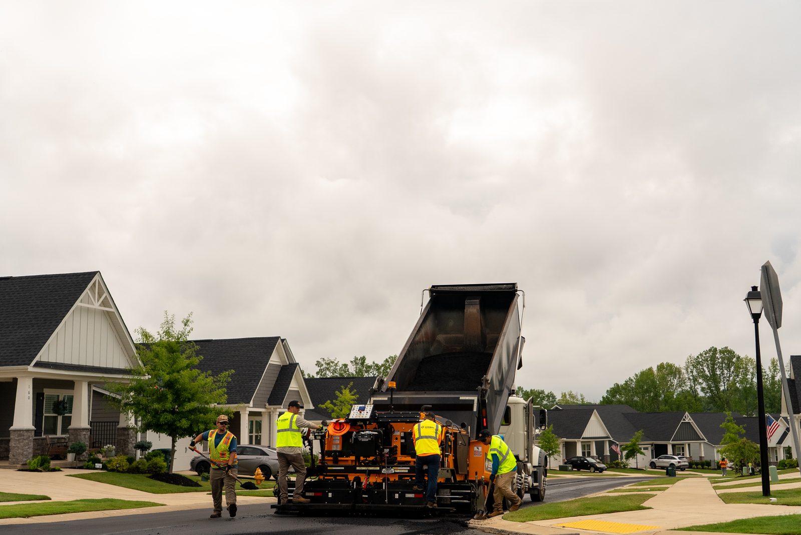 A dump truck raising its bed on a residential street under an overcast sky, new houses behind it