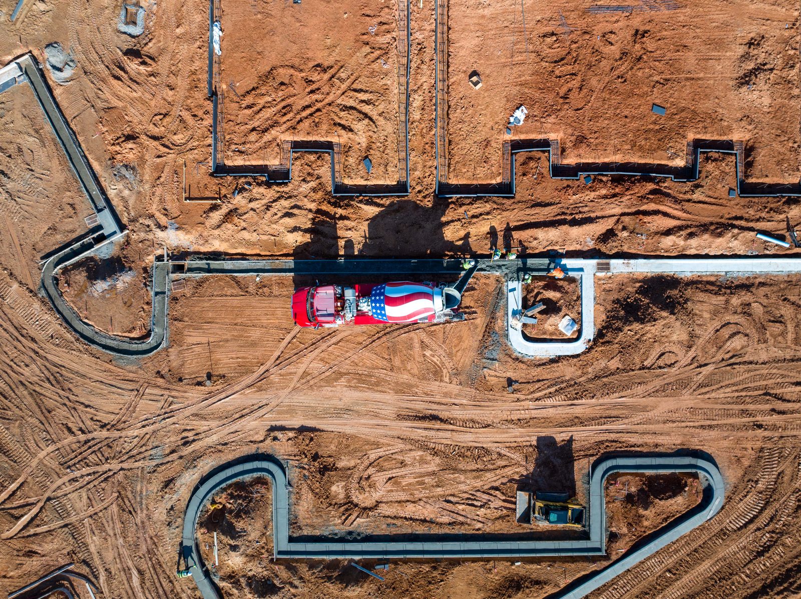 Overhead of a foundation under construction in red clay, formwork and pipe runs laid out, an American flag spread across the centre