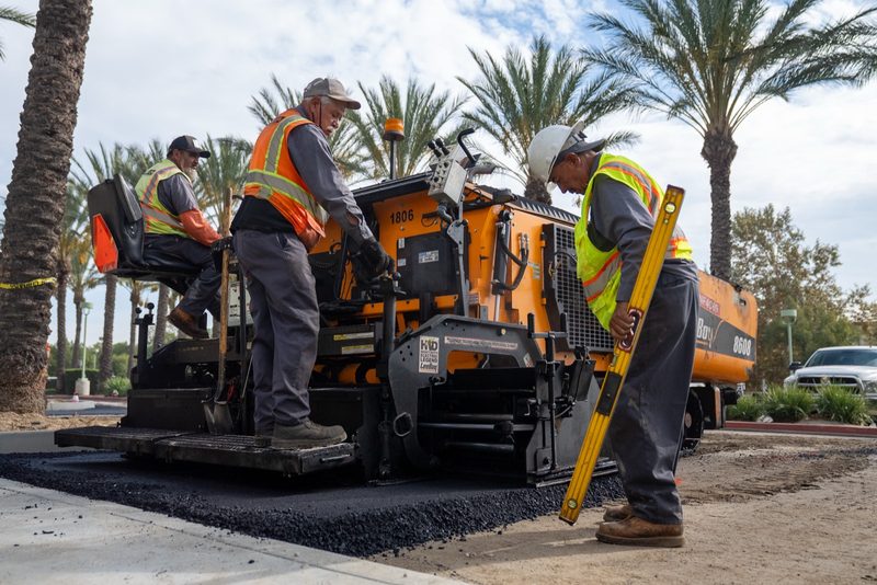 Crew setting grade behind a LeeBoy paver, palms along the kerb