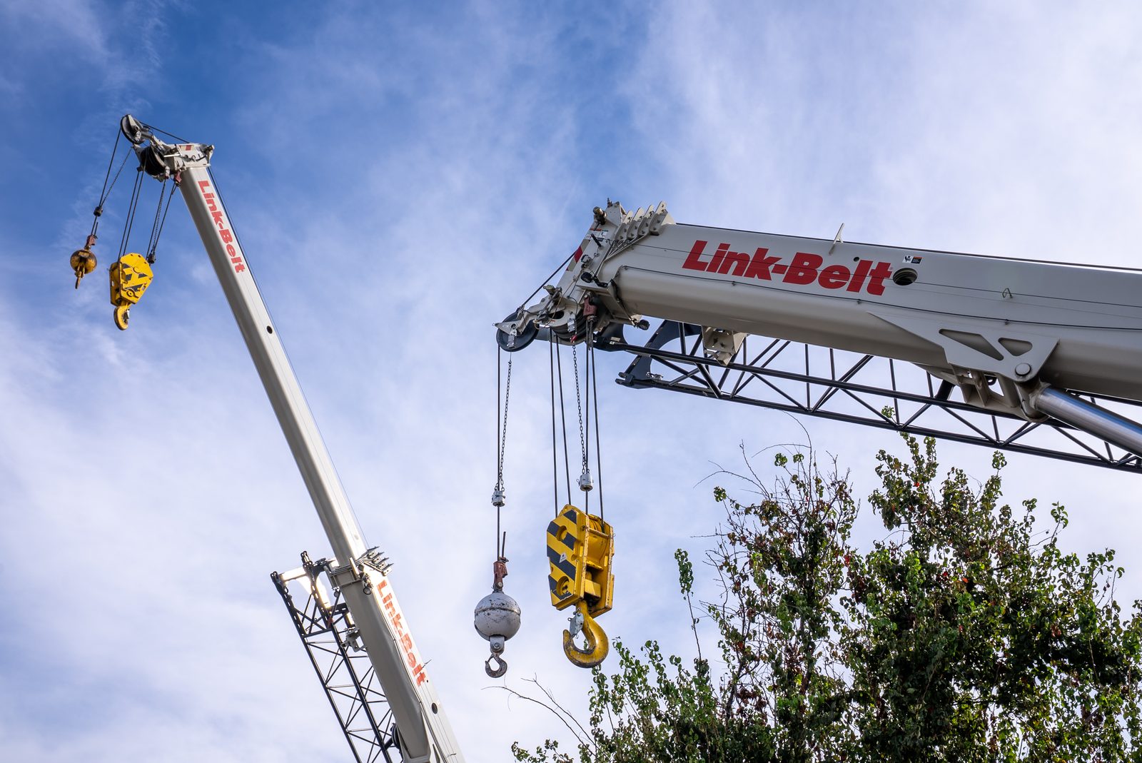 Two crane booms crossing against blue sky, a Link-Belt boom in the foreground with its rigging and hook blocks hanging