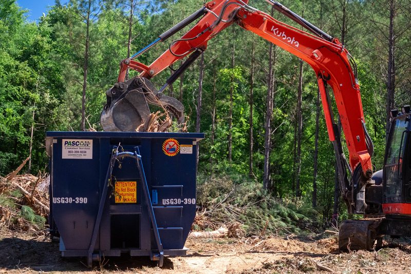 A grapple excavator feeding brush into a Pascon roll-off container