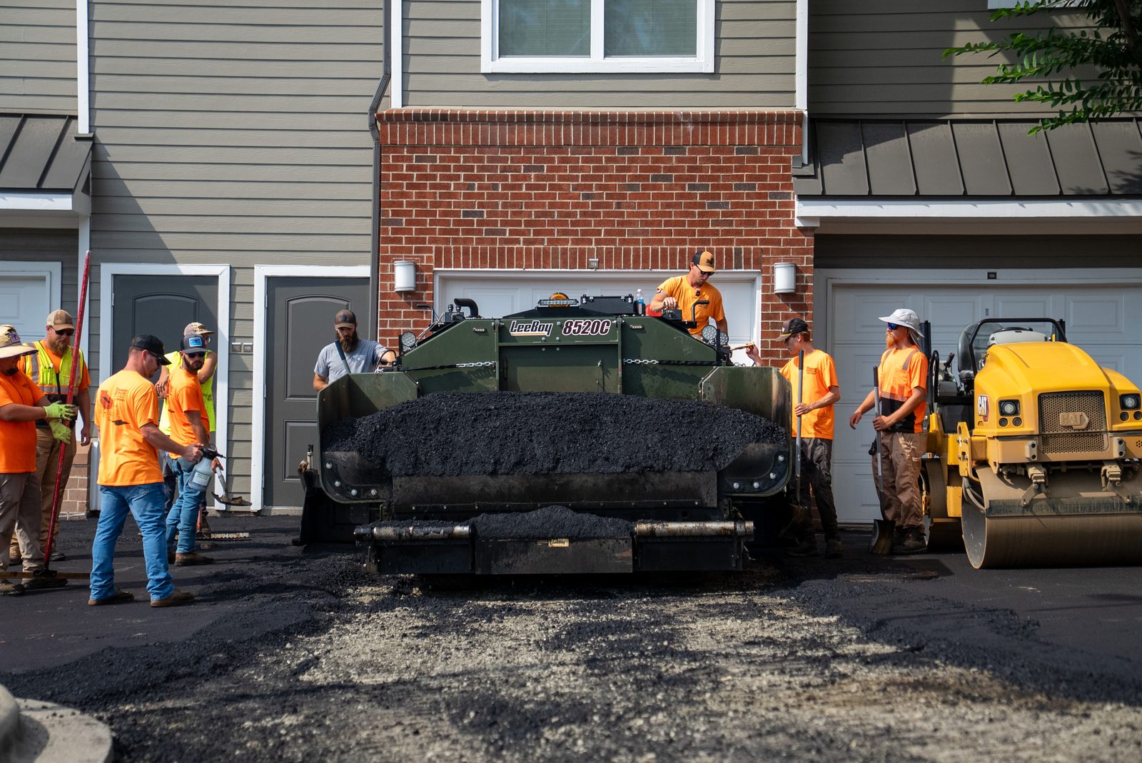 A Piedmont Paving crew in orange high-vis working a paver and roller across a fresh asphalt mat beside a brick building