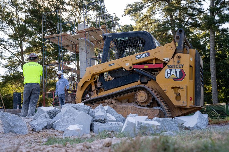 A CAT track loader working the rubble, crew watching from the bank