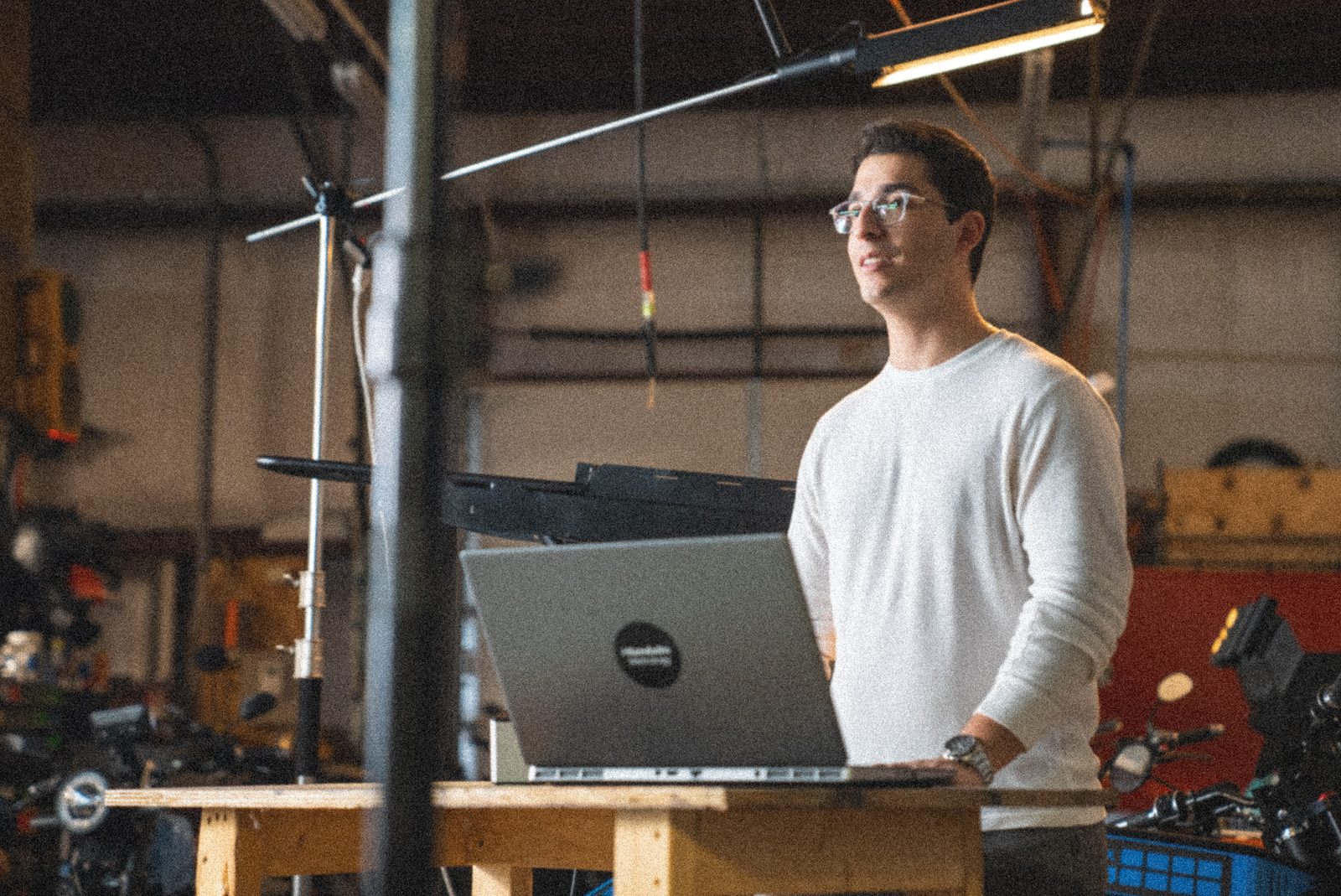A man in a workshop standing beside a laptop on a stand, equipment on the bench behind him