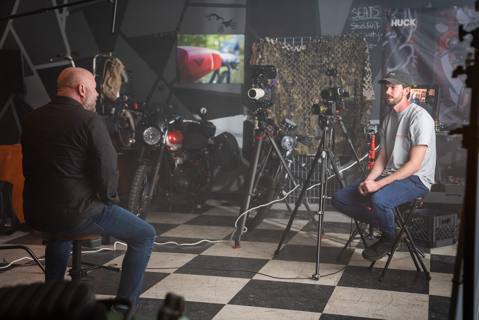 Wide on the interview set in the motorcycle shop, interviewer and subject facing off across the checkerboard floor
