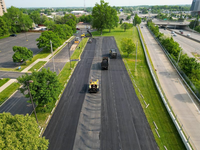 Drone down a divided street, the paving train mid-pull
