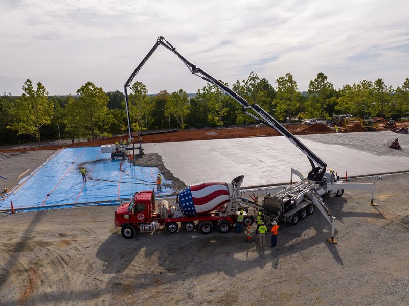Aerial on a slab pour, boom pump reaching across the deck to the mixer